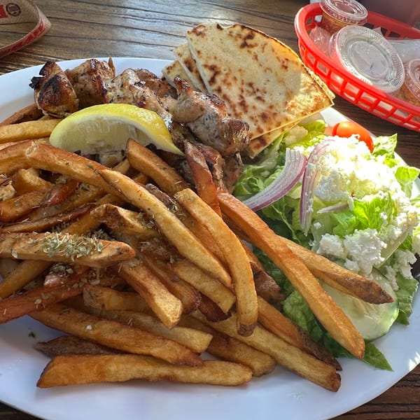 A plate of chicken souvlaki, seasoned fries, pita bread, and a Greek salad with feta cheese.