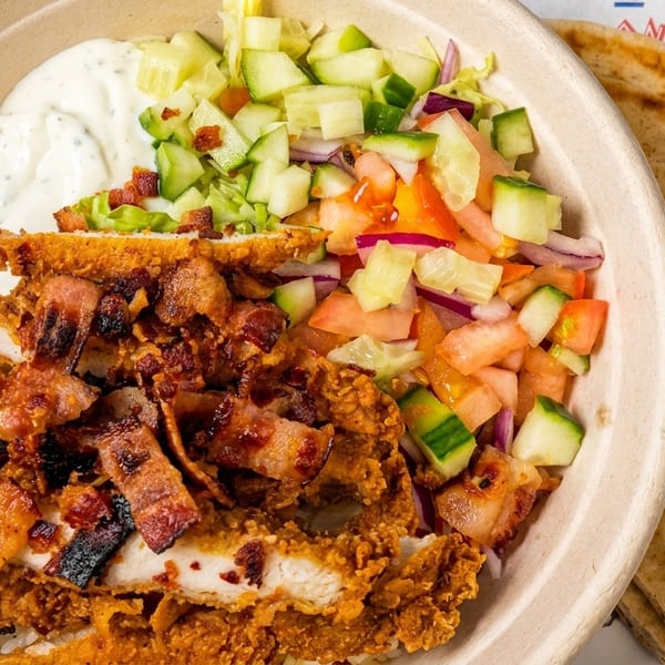 A Greek bowl with crispy chicken, bacon, rice, tzatziki, and a fresh cucumber and tomato salad.